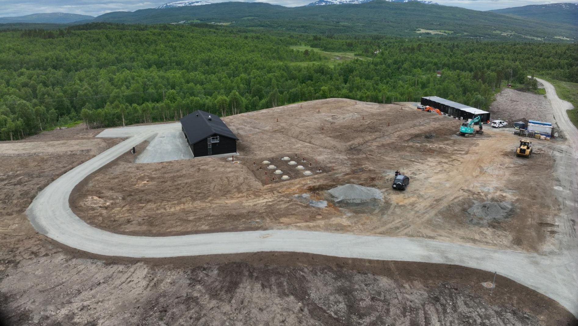Dronebilde av byggeplass med sort bygg, maskiner og anleggsvei omgitt av skog og fjell i bakgrunnen.
