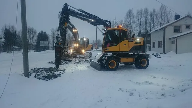 Gravemaskiner jobber i snøen utenfor bolighus med mørke trær i bakgrunnen.