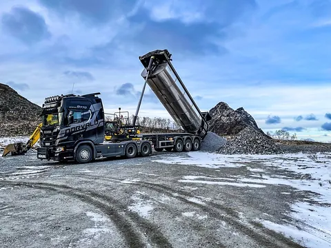 Tippbil losser grus foran en steinhaug i vinterlandskap med gravemaskin i bakgrunnen.