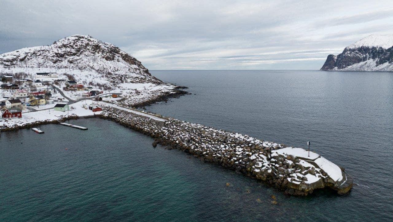 Molo med snø og store steiner strekker seg utover sjøen med fjell og småbåthavn i bakgrunnen.