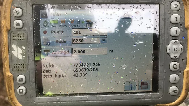 Nærbilde av en våt GPS-enhet fra Topcon med koordinatdata på skjermen.
