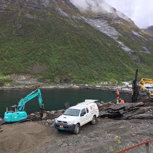 Gravemaskiner og servicebil i arbeid ved fjord, med grønt fjell i bakgrunnen og arbeidere på stedet.