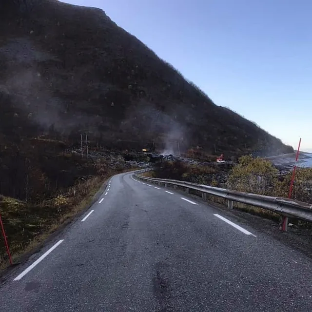 Asfaltert vei ved fjord og fjell, med røyk fra anleggsarbeid synlig i det fjerne.