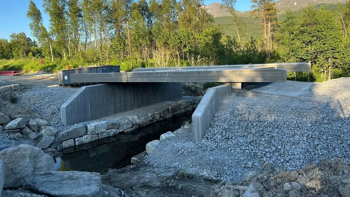 Elveforbygning med naturstein og støpt betongbru under montering i solfylt skogsområde.
