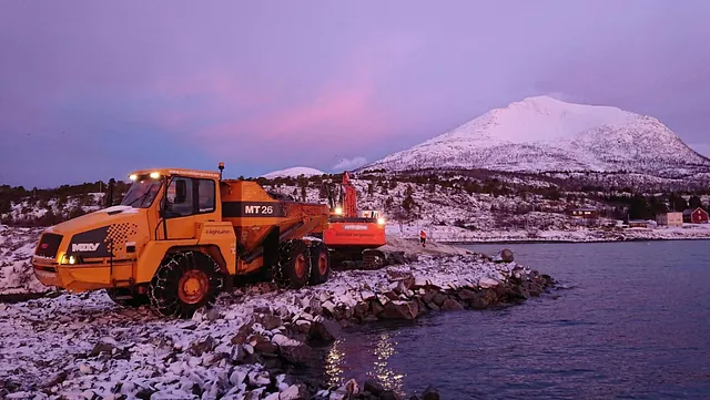 Dumper og gravemaskin arbeider på snødekt steinfylling ved sjøen, med fjell i rosa kveldssol bak.