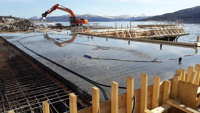 Betongdekke under bygging ved sjøen med armering og gravemaskin i bakgrunnen.