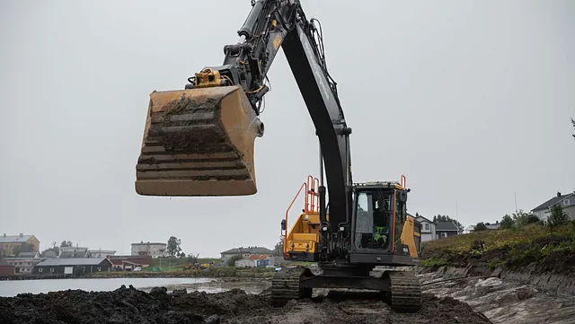 Gravemaskin med hevet skuff står ved vannkanten i et vått og gjørmete landskap.