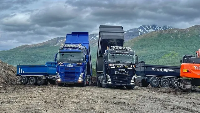 To lastebiler tipper masser på en anleggsplass foran grønne fjell og grå himmel.