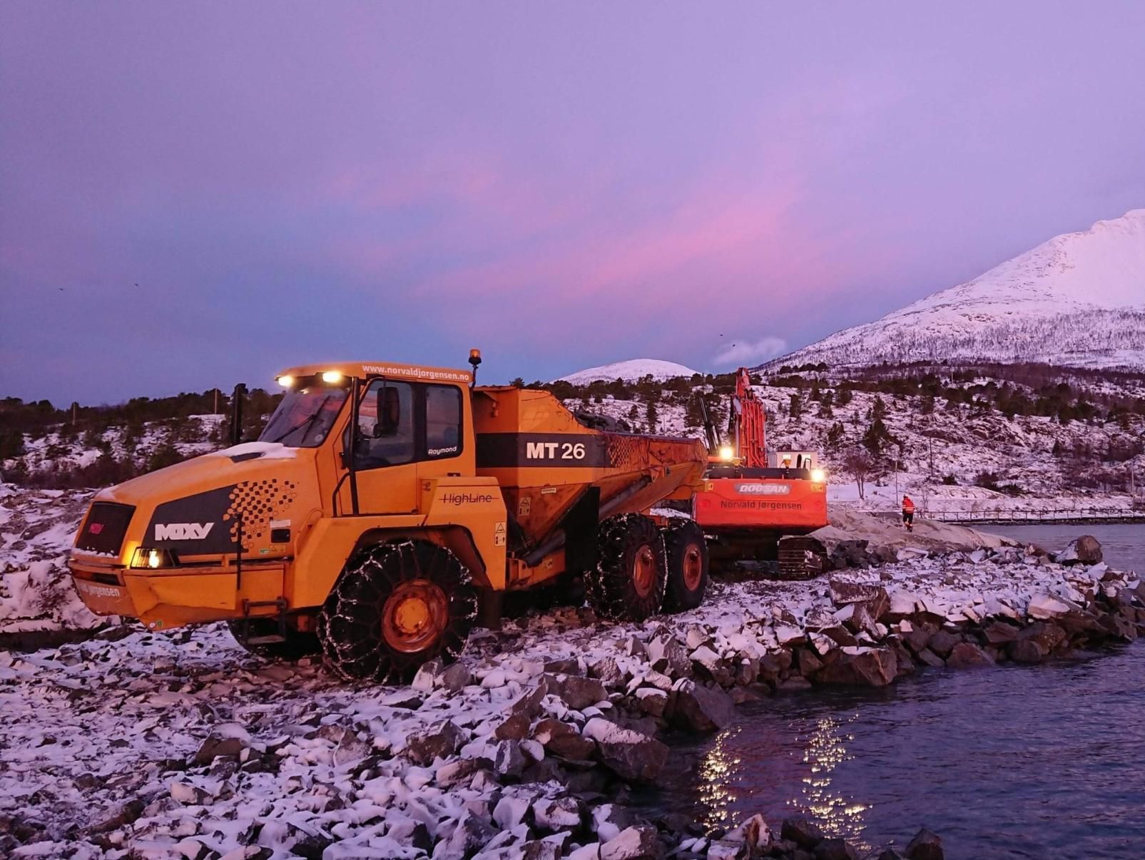 Dumper og gravemaskin arbeider på snødekt steinfylling ved sjøen, med fjell i rosa kveldssol bak.