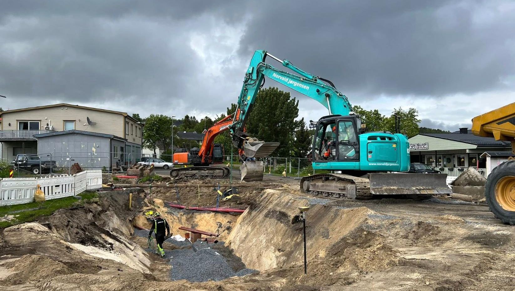 Gravemaskin jobber i en dyp byggegrop ved bolighus, med mørke regnskyer over.