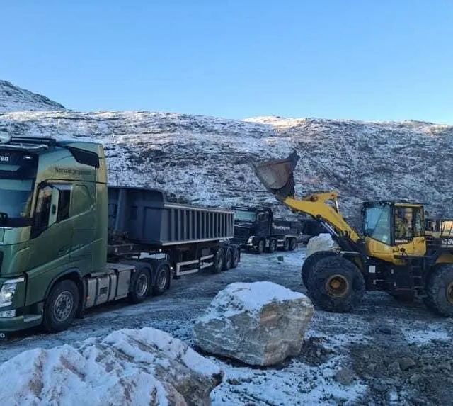 Gul hjullaster laster masser i en grønn lastebil med henger i et snødekt fjellområde.