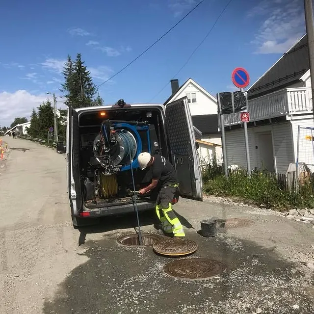 Arbeider renser kumlokk med slange fra servicebil i boligområde med grusvei.